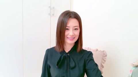 Video of a smiling East Asian woman with straight, shoulder-length brown hair, wearing a black satin blouse with a bow tie, seated in a room with white walls and a beige cushion.