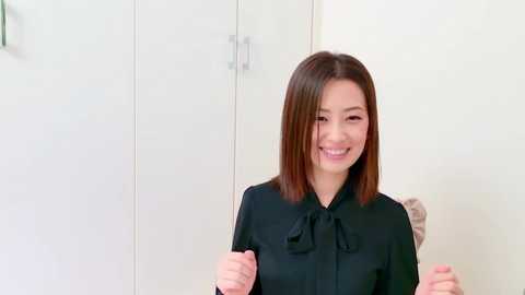 Video of a smiling Asian woman with straight, shoulder-length brown hair, wearing a black blouse with a bow, standing in a minimalistic, white room with closed cabinets in the background.