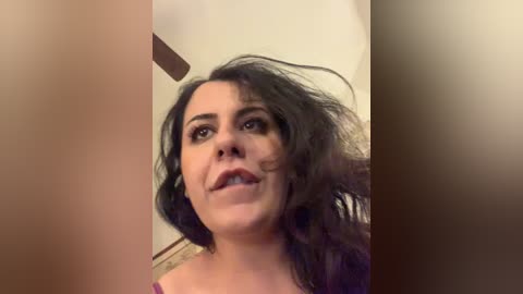 Video of a woman with dark, wavy hair and fair skin, wearing a purple top, standing indoors with blurred background. She has a neutral expression, and the image has a soft, warm tone.