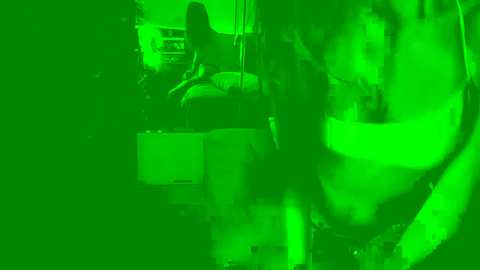 A green-tinted video of a nude woman with dark hair, seated on a chair, with a blurry, partially obscured male figure in the background. The image is dimly lit, creating a surreal, slightly distorted atmosphere.