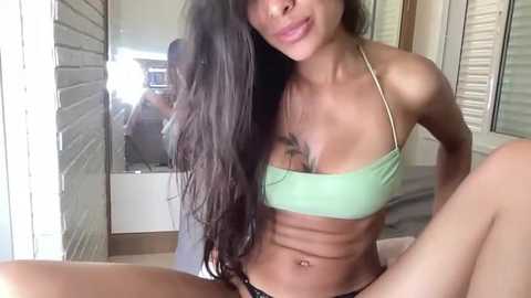 Video of a fit, light-skinned woman with long black hair, wearing a green bikini top, sitting on a bed, with a mirror reflecting a room with white blinds and a wooden floor.