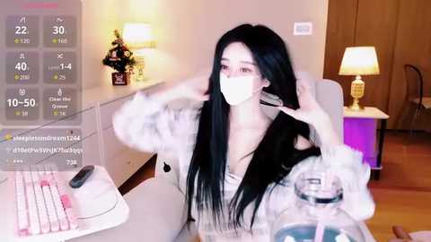 Media: Video of an Asian woman with long black hair, wearing a white mask, sitting at a table, adjusting her hair. Background features a dimly lit room with a lamp, wooden furniture, and a partially visible person in the background.