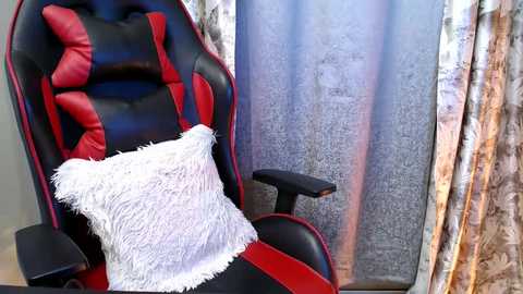 Video of a red and black gaming chair with a white fluffy pillow, situated in a room with floral-patterned curtains.