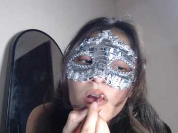 Video of an Asian woman with long black hair, wearing a silver masquerade mask with intricate designs, touching her lips. She is indoors, in a room with a mirror and a dark background.