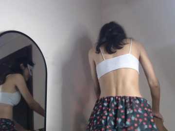 Video of a slender woman with shoulder-length black hair, wearing a white sports bra and floral skirt, standing in a dimly lit room, facing a mirror.