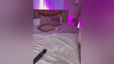 A dimly-lit bedroom with a plush, light-colored bed, adorned with pillows and a red heart banner, features a vibrator on the bedspread. Soft purple lighting enhances the room's cozy atmosphere.