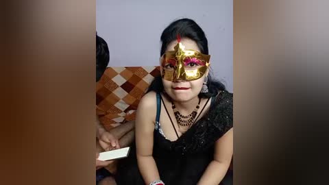 Video of a woman in a black dress, adorned with a golden masquerade mask and intricate black necklace, seated on a checkered-patterned cushion.