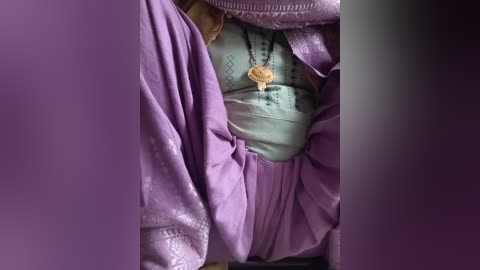 Video of a person's chest and upper abdomen, wearing a light green shirt with intricate embroidery. A purple, flowy, textured robe drapes over the shirt, adorned with a large, golden pendant. The background is blurred, with purple tones dominating the frame.