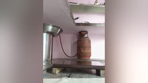 A video of a small, unfinished kitchen space with a silver gas stove and a rusted, red \"Indane\" gas cylinder propped against a light pink wall. The floor is a mixture of concrete and dirt, and the ceiling is unfinished.
