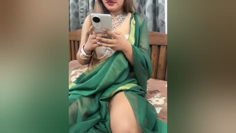 Video of a South Asian woman with medium skin tone, wearing a green saree with a sheer blouse, sitting on a bed, taking a selfie.