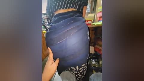 Media: Video of a woman's lower back and buttocks, wearing a blue floral-patterned skirt, with a hand pulling up the skirt to reveal her round, firm buttocks. Background shows a cluttered room with kitchen items and a red container.