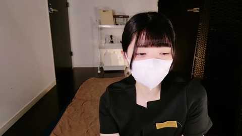 Video of an Asian woman with short black hair and a white surgical mask, wearing a black scrub top. She stands indoors on a wooden floor, with a beige towel and a white shelving unit in the background.