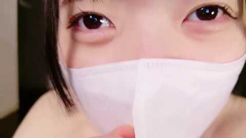 A close-up video of an Asian woman with fair skin, wearing a white medical mask that covers her mouth and nose. Her large, dark brown eyes are visible, gazing intently. The background is blurred, focusing on her face.