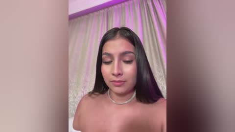 Media: A video of a young woman with long, straight black hair, wearing a silver necklace, standing against a backdrop of light beige curtains with purple lighting.