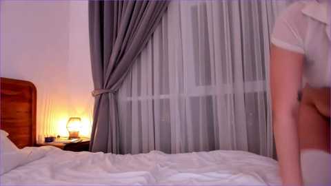 Media: Video of a dimly lit bedroom with a bed, a wooden headboard, white sheets, and gray curtains partially drawn. A lamp with a glass shade is on a wooden nightstand.