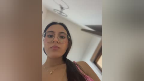 Video of a young woman with long dark hair, wearing glasses and a pink lace bra, standing in a dimly lit room with a ceiling fan.