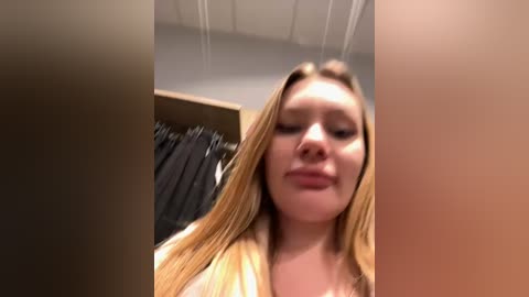 Media: Video of a young, fair-skinned woman with long, straight blonde hair, wearing a light-colored top, looking down and smiling slightly, standing in a dimly lit, cluttered room with a wooden wardrobe and hanging clothes in the background.