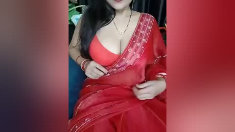 Media: Video of a woman with long black hair, wearing a red saree with a deep neckline, revealing cleavage. She sits against a black background, adorned with gold jewelry.