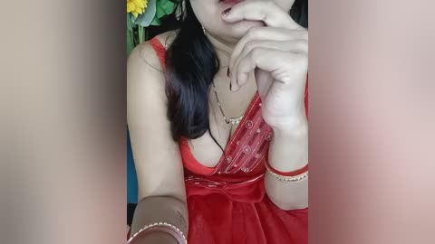 Media: Video of an Asian woman with long black hair, wearing a red lace bralette, gold necklace, and a bracelet, biting her finger, against a background of flowers and a blue wall.