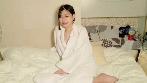 Media: Video of a young Asian woman with short black hair, smiling while sitting on a bed in a white bathrobe, surrounded by beige and brown patterned pillows, in a simple, modern bedroom.