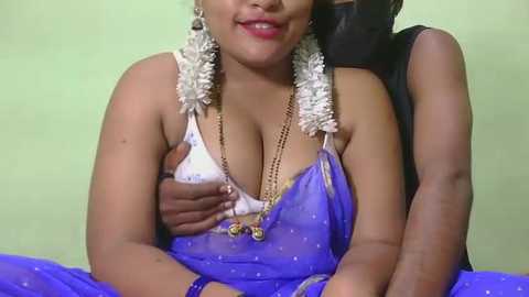 Video of a woman with medium skin tone wearing a blue saree with gold trim, white flowers in her hair, and gold jewelry, sitting on a bed, with a man's arm around her.