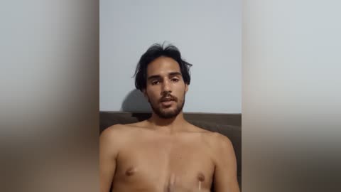 Video of a shirtless, middle-aged man with medium-dark skin tone, short black hair, and a trimmed beard. He stands in front of a white wall with a shadow on the left side, revealing a rectangular shape.