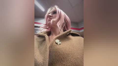 Video of a woman with pastel pink hair, wearing a brown coat with a button, standing in a fluorescent-lit room with red and white striped walls.