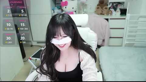 Media: Video of a smiling Asian woman with long black hair, wearing a white blindfold, black top, and white jacket, sitting on a white chair in a room with a white bed, pink teddy bear, and a pink chair.