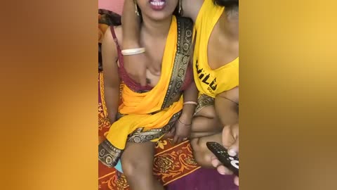 Media: A video of two women in vibrant yellow sarees with gold trim, one showing cleavage, sitting on a red and gold patterned cushion. The background features a blurred room with a yellow gradient.