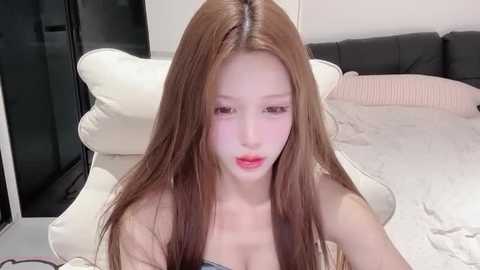 Media: Video of an East Asian woman with long, straight brown hair, fair skin, and light makeup. She wears a strapless top, sitting on a bed with white sheets and pillows, in a modern, minimalist bedroom.