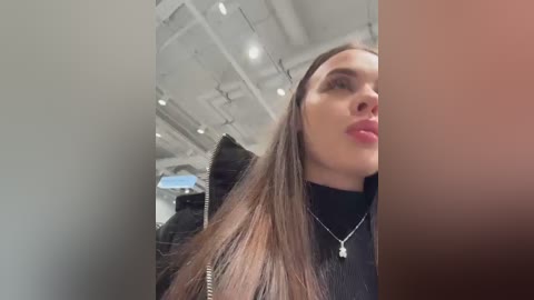 Media: A video captures a young woman with long, straight brown hair, wearing a black jacket and silver necklace, inside a modern, well-lit room with exposed ceiling beams.