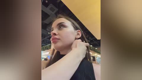 Media: Video of a young woman with fair skin, long brown hair, and brown eyes, wearing a black top, gazing upward, standing in a brightly lit indoor space with a yellow triangular overlay.