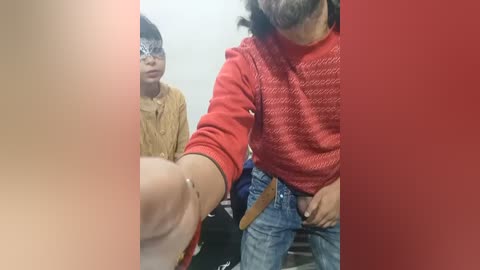 Video of a man in a red sweater, leaning forward, and a woman in a yellow top and mask, seated in a room with red vertical filter overlay.