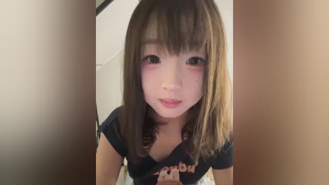 Media: A young Asian woman with straight, shoulder-length brown hair and light skin, wearing a black Hooters t-shirt, blushing, and smiling. Indoor background with blurred beige and white tones.