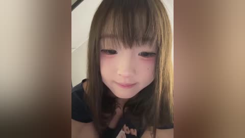 Media: Video of a young Asian woman with fair skin, straight brown hair, and a light makeup look, leaning forward, wearing a dark top. Background is blurred and neutral-colored.