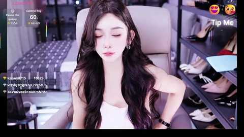 A video of a young Asian woman with long black hair, wearing a white dress, seated in a modern room with a glass table and shelves of shoes.