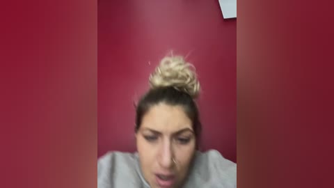 Media: Video of a young woman with a blonde bun, wearing a grey t-shirt, against a red wall, looking down, with a blurred, reddish background.