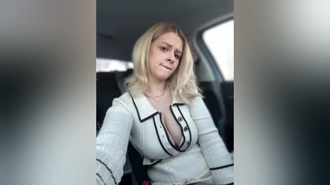 Video of a blonde woman with shoulder-length hair, wearing a white cardigan with black buttons, inside a car. She has a neutral expression and is seated in the driver's seat.