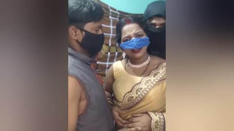 Media: Video of three individuals in a crowd, wearing face masks, one woman in a yellow sari with a blue mask, a man in a sleeveless shirt, and another person partially visible behind them.