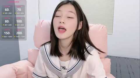 Media: Video of a young Asian woman with straight black hair, closed eyes, and slightly open mouth, wearing a white sailor school uniform, lying on a pink bed in a minimalistic room.