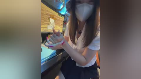Media: Video of a young woman with long brown hair and fair skin wearing a white face mask, white blouse, and black skirt, playing with a white stuffed animal on a countertop.