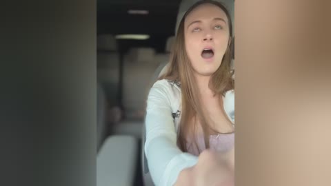 Video of a young Caucasian woman with long brown hair, wearing a grey beanie and white top, screaming in a car, blurry background.