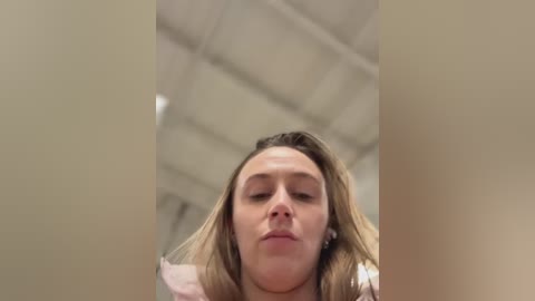 A video of a young woman with light skin, long blonde hair, and closed eyes, captured from a low angle, with a beige ceiling and industrial lighting in the background.