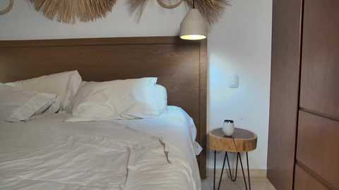 Video of a minimalist bedroom with a wooden headboard, white bedding, beige pouf, and a round wooden side table with a white vase. Decorative woven wall hangings and a modern pendant light above.