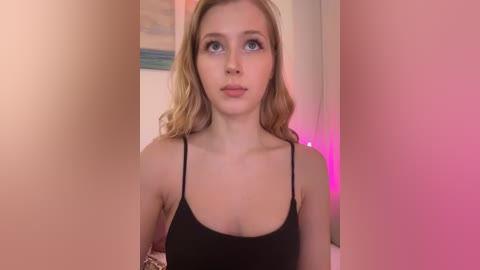 Media: Video of a young woman with fair skin and long, wavy blonde hair, wearing a black spaghetti-strap top, standing against a pink backdrop.