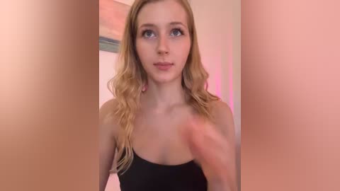 Media: Video of a fair-skinned, blonde-haired young woman with a slender build, wearing a black strapless top, looking at the camera with a neutral expression. Background shows blurred beige and pink walls.
