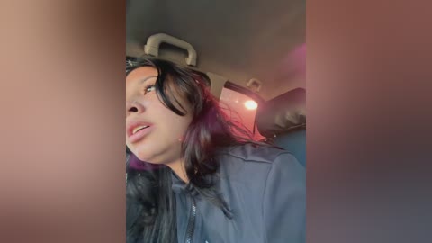 Media: Video of a woman with long, dark hair and fair skin, wearing a black leather jacket, looking out of a car window, with a blurred, orange-tinted background.