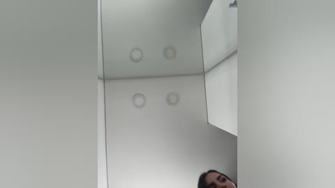 Video of a minimalist, white room with a reflective ceiling featuring two circular light fixtures, partially obscured by a person's head and shoulders.