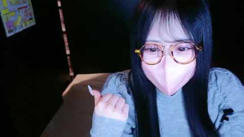 Video of an East Asian woman with long black hair, wearing a surgical mask and glasses, seated at a dimly-lit table, gesturing with her right hand.