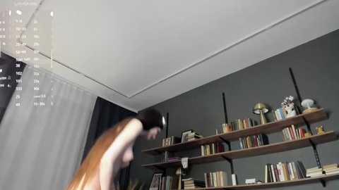 A video of a naked woman with a slender build and short dark hair, captured mid-motion, standing in a dimly lit room with gray walls and bookshelves.